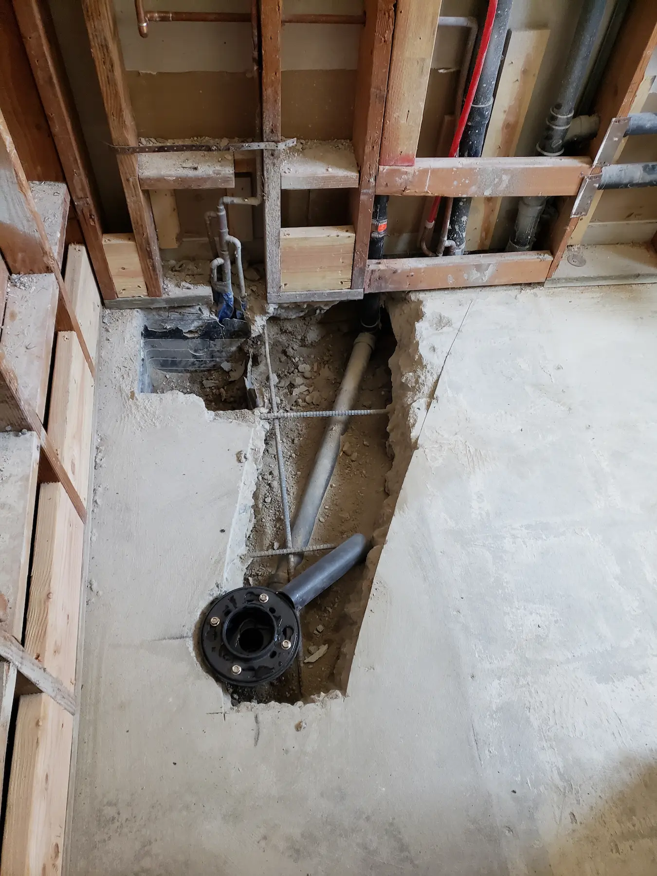 Concrete floor cut open to expose plumbing pipes and wooden framing behind a wall.